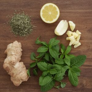 A close-up image showing ingredients for green tea preparation: a halved lemon, fresh ginger slices, mint leaves, dried green tea leaves, and a piece of ginger root arranged on a wooden surface.